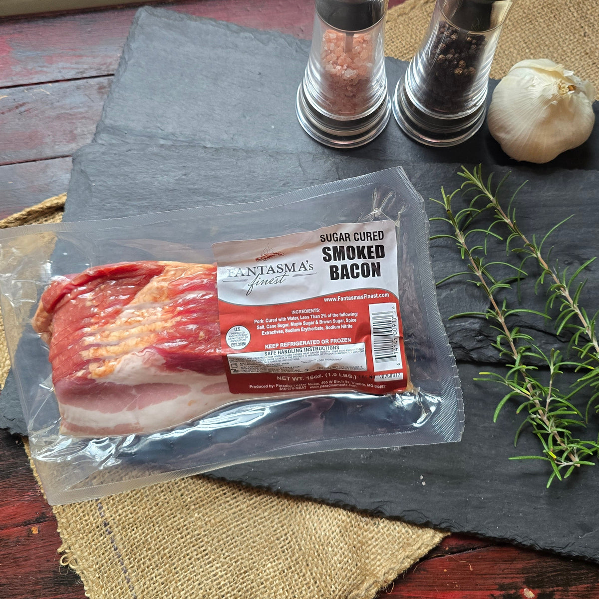 A vacuum-sealed package of smoked bacon lies on a dark slate board, styled with a sprig of rosemary, a garlic bulb, and two salt and pepper mills on a rustic red wooden surface.