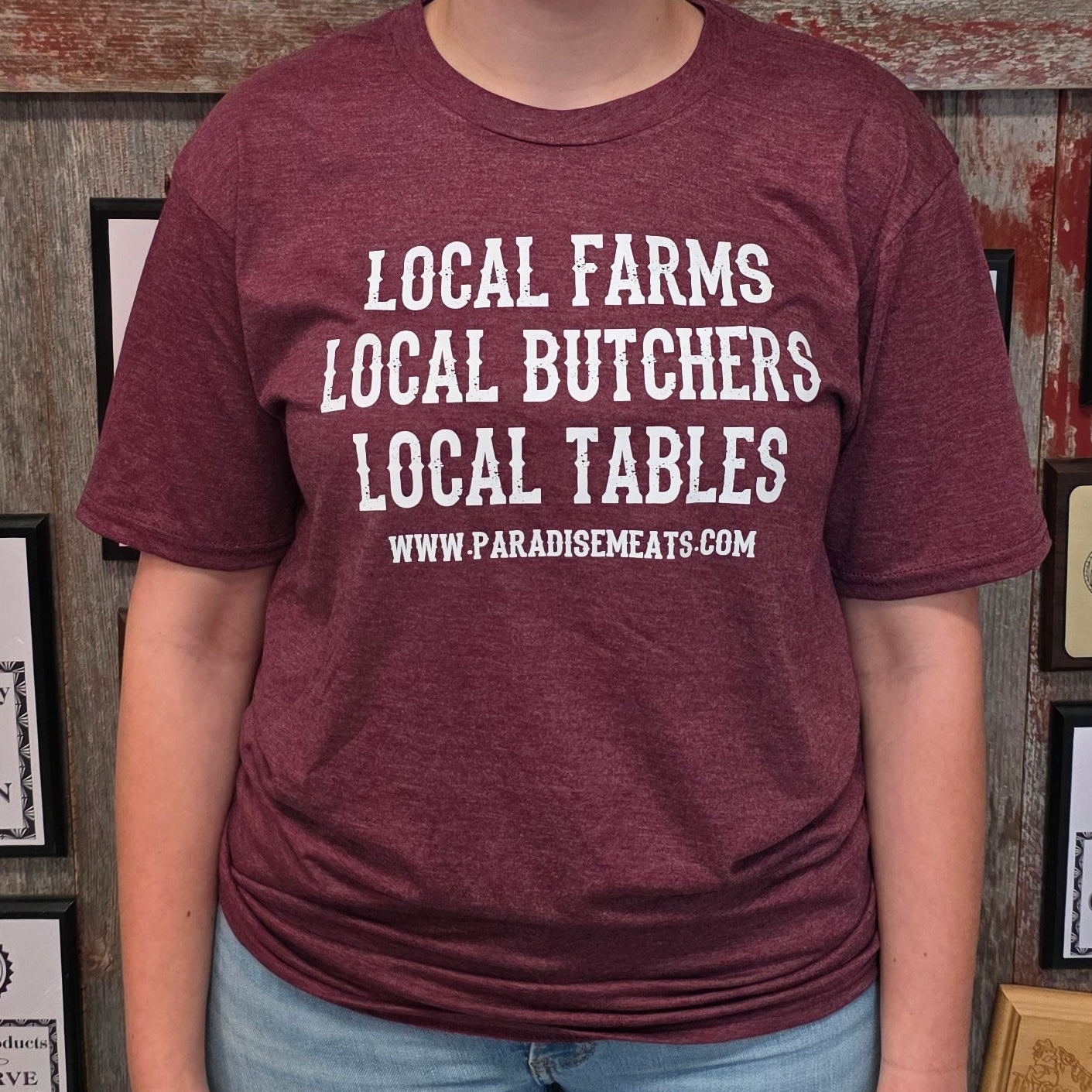 A woman stands in front of a rustic wooden wall covered in framed awards and certificates, wearing a heathered maroon t-shirt with white text that reads 'LOCAL FARMS LOCAL BUTCHERS LOCAL TABLES' and light blue jeans.