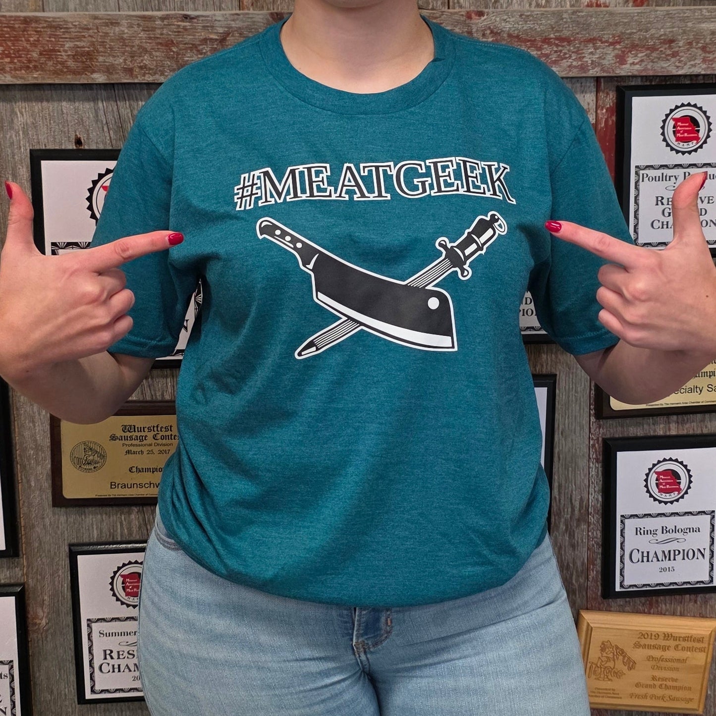 A woman stands in front of a wall covered with championship plaques, wearing a teal graphic t-shirt that says '#SMILE WITH A KNOT' with a black bow and arrow design, paired with light blue jeans and dark boots.