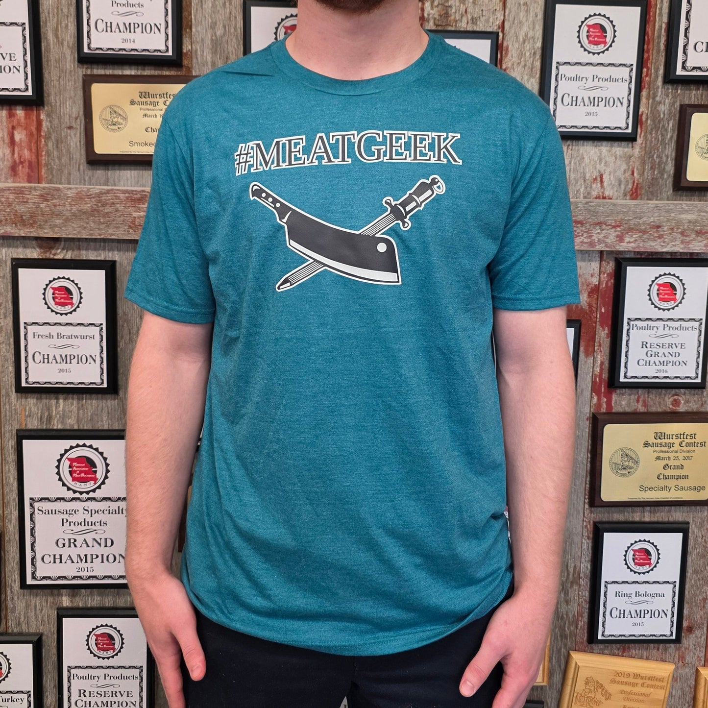 A man with blond hair and a beard wearing a teal graphic t-shirt with a whale and sword design and the text '#SMEATGEEK', standing in front of a wall covered in framed awards and certificates.