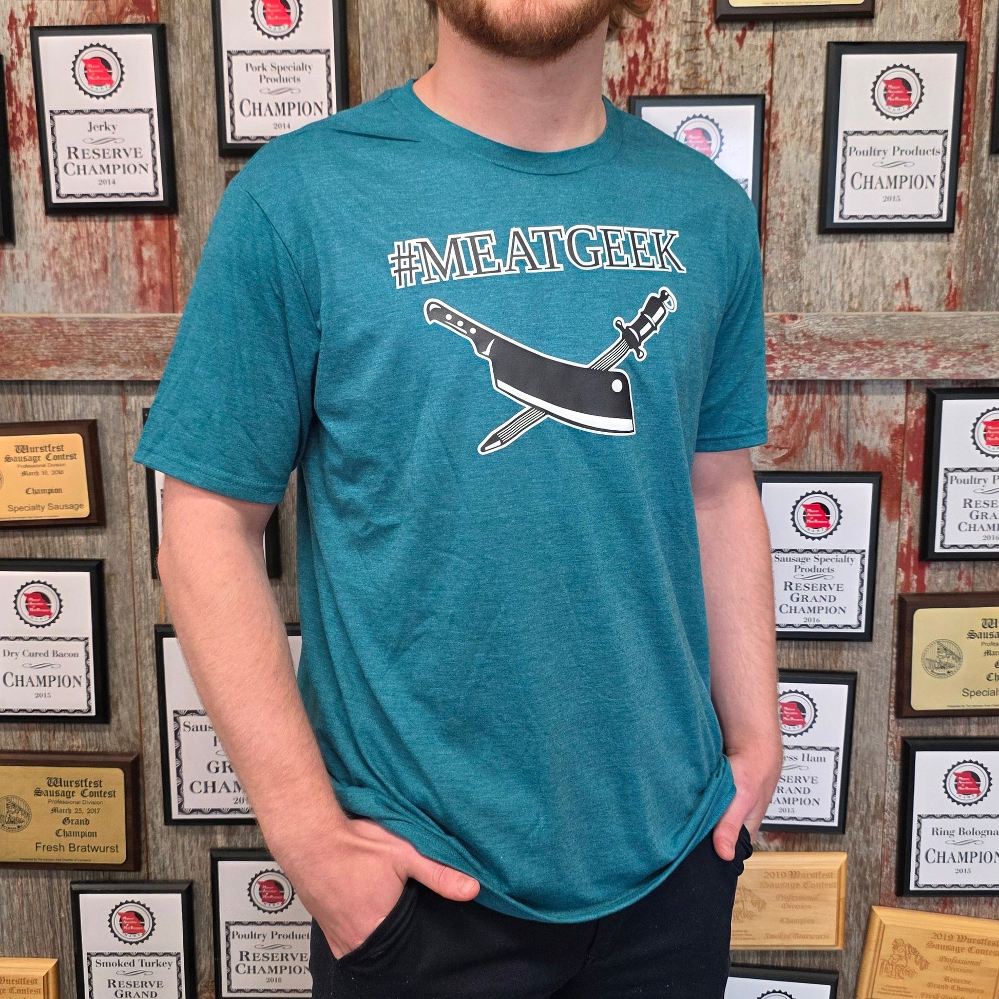 A smiling young man with red hair and a beard stands in front of a wall covered in framed awards and plaques, wearing a teal graphic t-shirt that reads 'FANTASTIC GEEK' with a cartoon character, and dark pants.