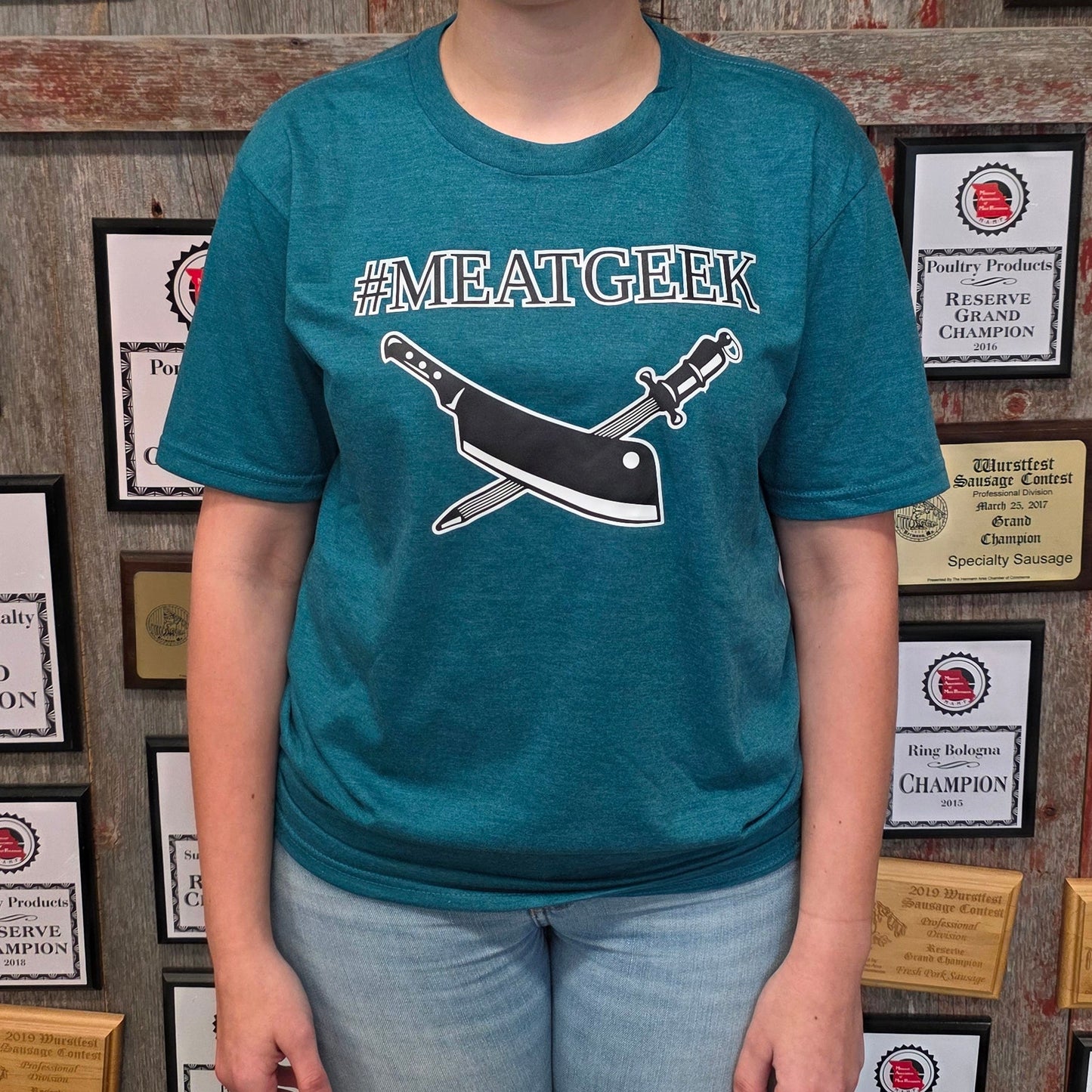 A woman stands in front of a wall covered in framed awards and championships, wearing a teal graphic t-shirt with a whale and sword design and the text '#MREATGEEK', paired with light blue jeans and dark rubber boots.