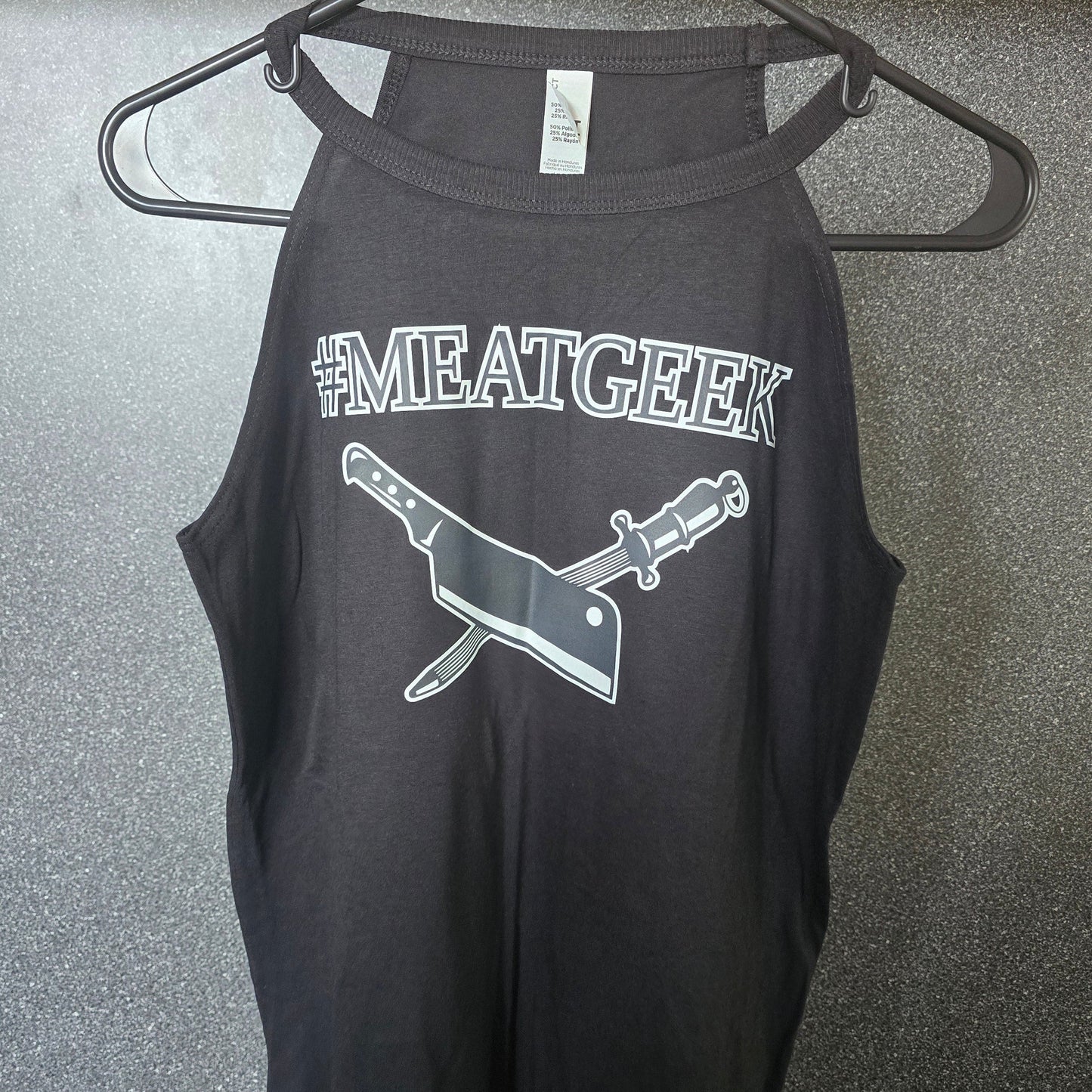 A black sleeveless tank top hanging on a black metal hanger against a textured gray wall. The shirt features a white graphic print of a cleaver and knife with the hashtag '#MEATGEEK' above it.