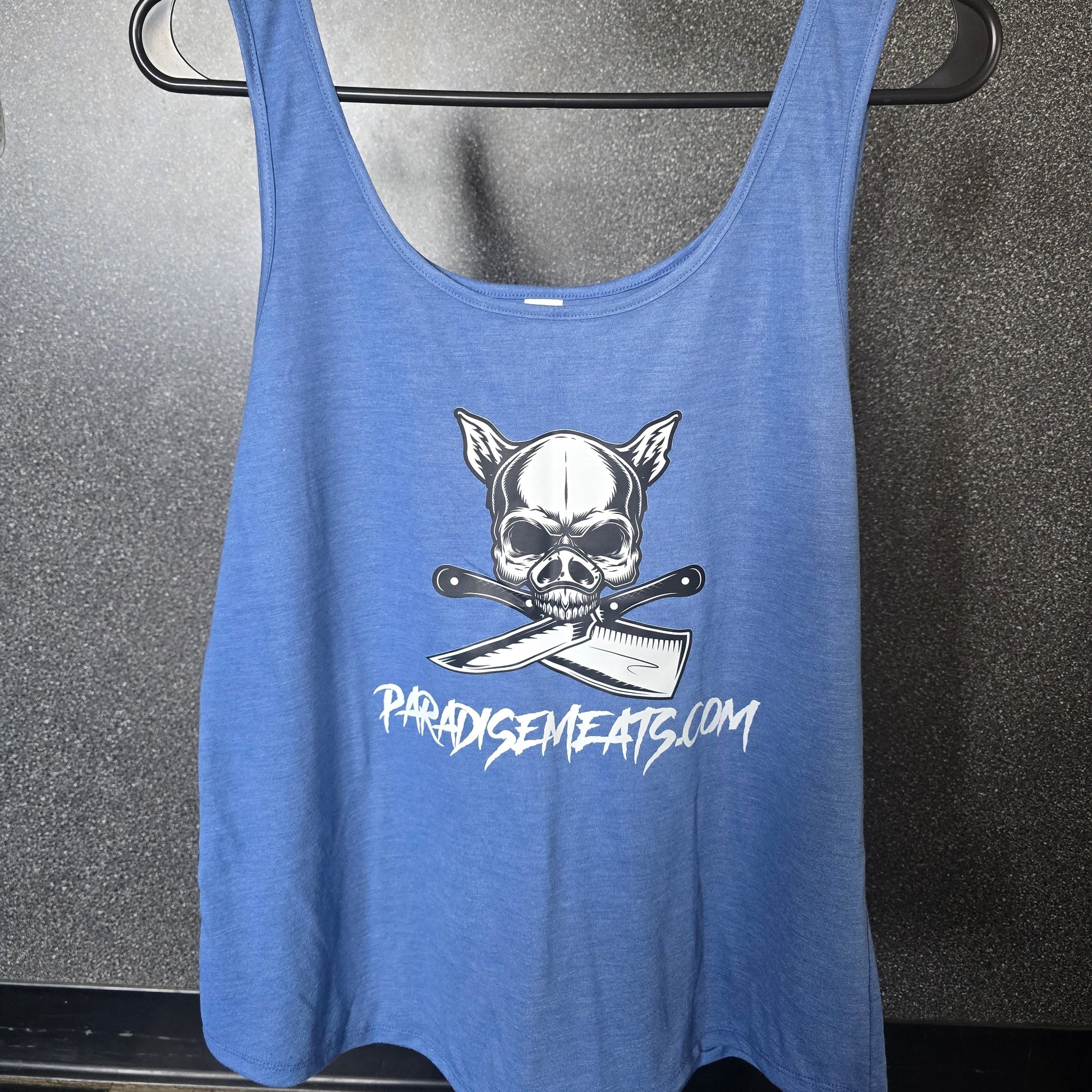 A blue tank top with a white graphic of a skull wearing a chef's hat over crossed knives, with the text 'PARODYCEMENTS.COM' printed below, hanging on a black clothes hanger against a dark gray floor.