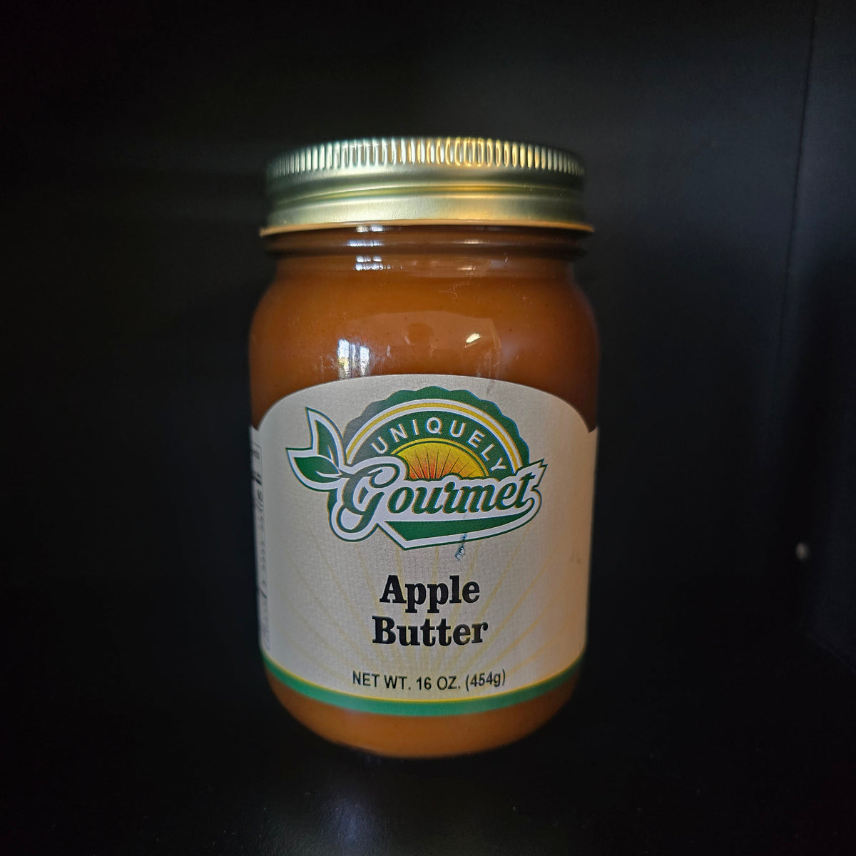 A 16 oz glass jar of Gourmet Apple Butter with a gold metal lid and a white label featuring the 'Uniquely Gourmet' logo, set against a dark background.