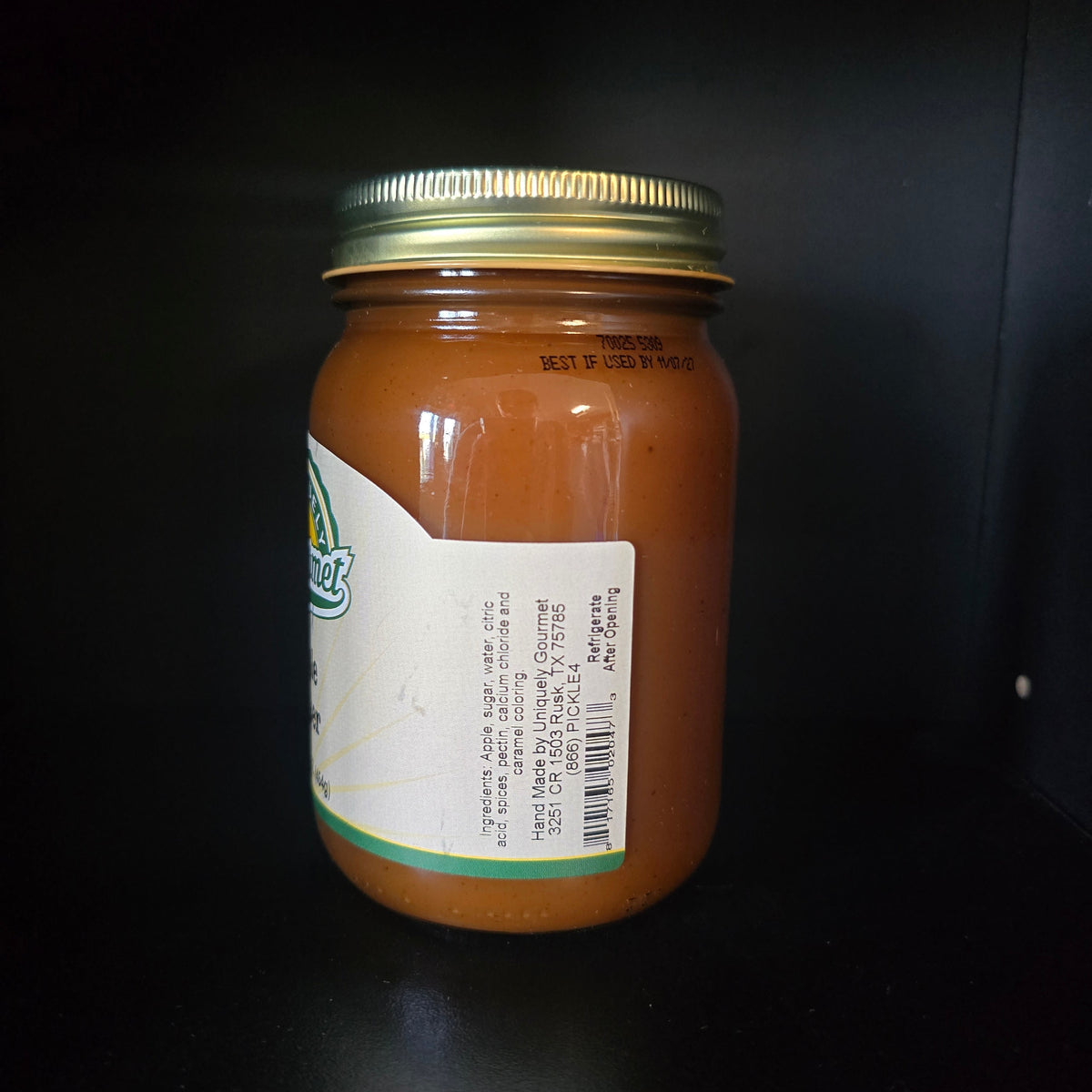 A glass jar filled with orange-brown sauce or preserve, sealed with a gold metal lid and featuring a white label with green accents, photographed against a dark background.