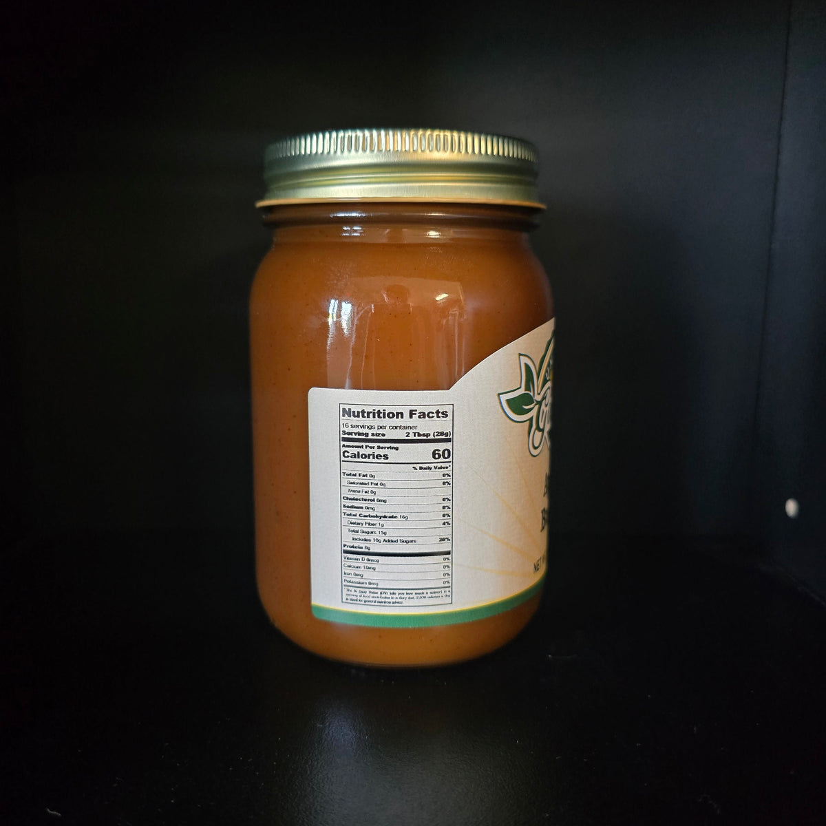 A glass jar filled with orange-brown liquid, sealed with a gold metal lid and featuring a cream label with green leaf graphics, photographed against a dark background.