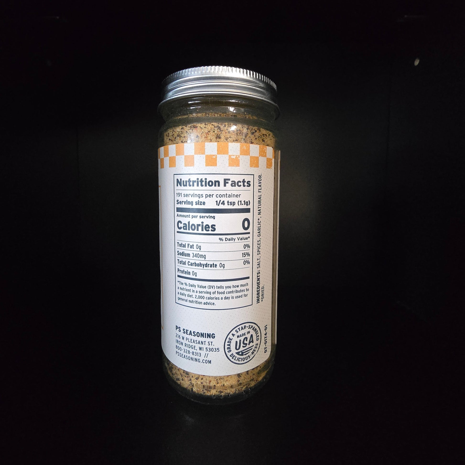 A glass jar of seasoning with a white label and silver lid, standing on a black reflective surface against a black background.