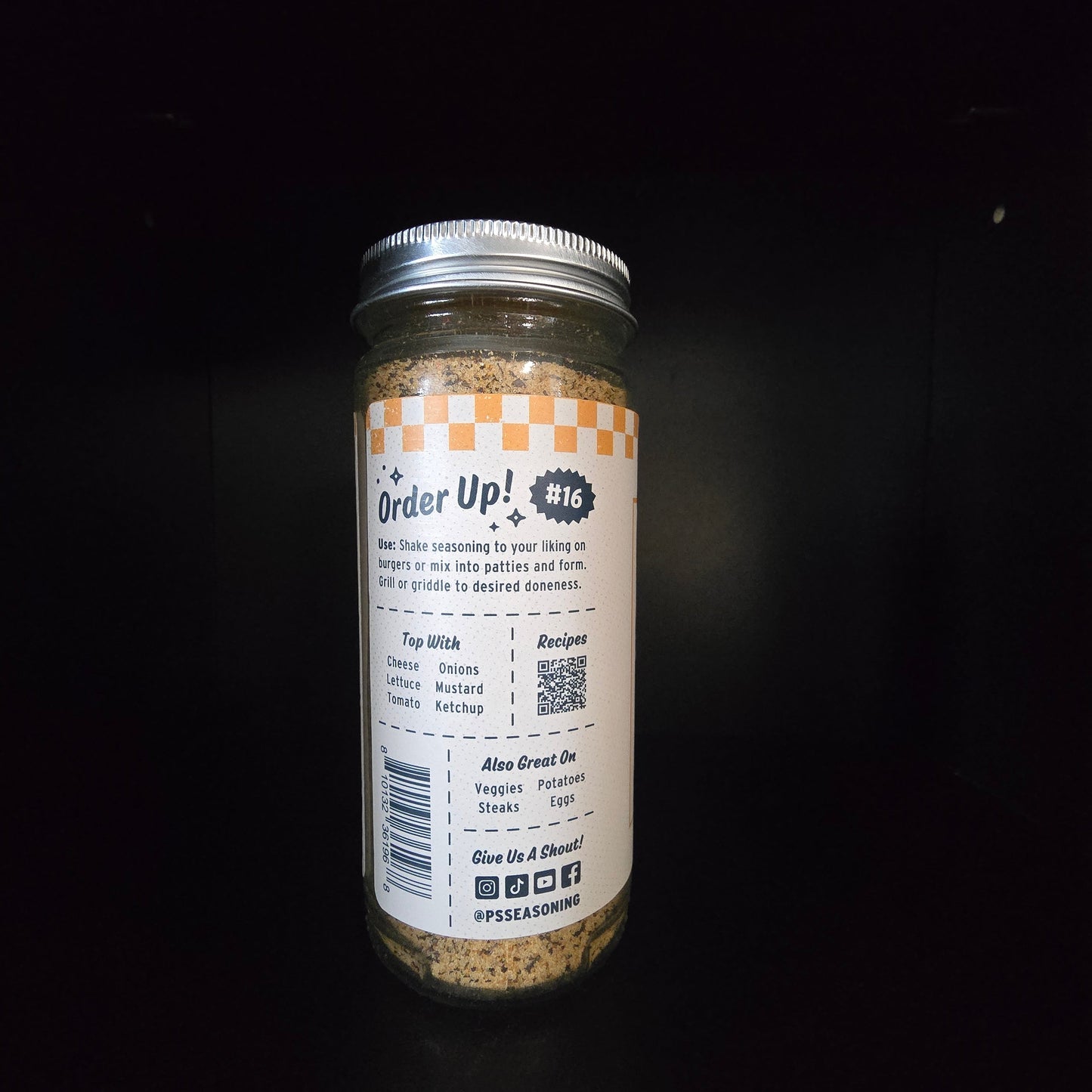A glass jar of seasoning mix featuring a white label with orange accents and a silver metal lid, set against a dark background.