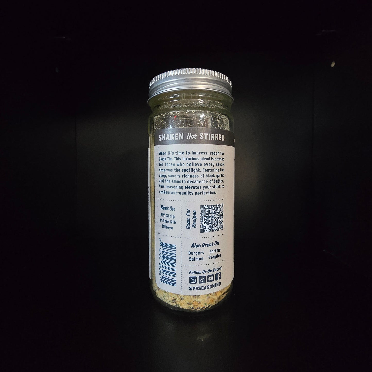 A glass jar of seasoning with a silver metal lid, shown from the back against a dark background.