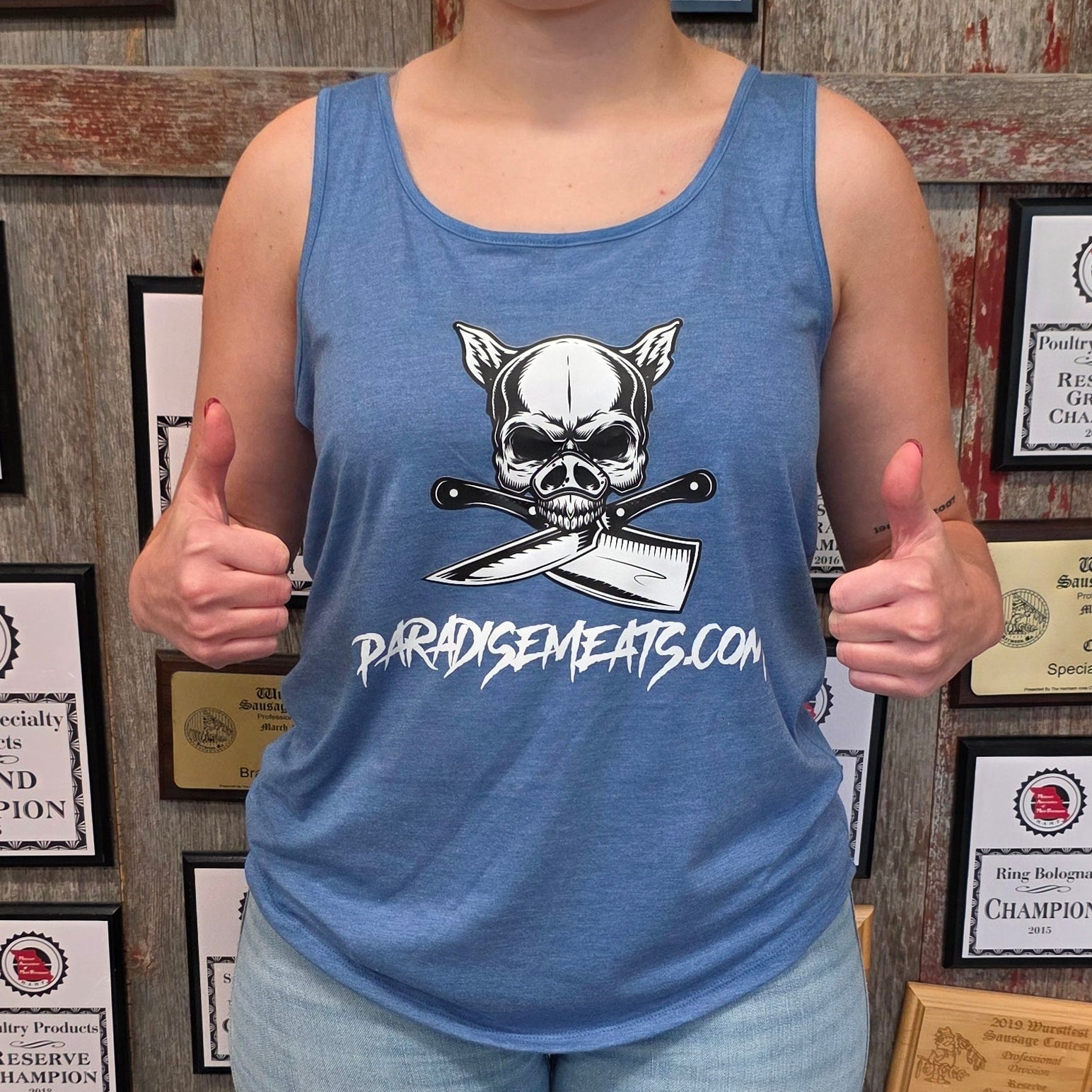 A woman stands in front of a wall covered in framed championship certificates, wearing a blue tank top with a skull and crossbones graphic and the text 'PROBLEMASKERS.COM', paired with light blue jeans and dark green rain boots.