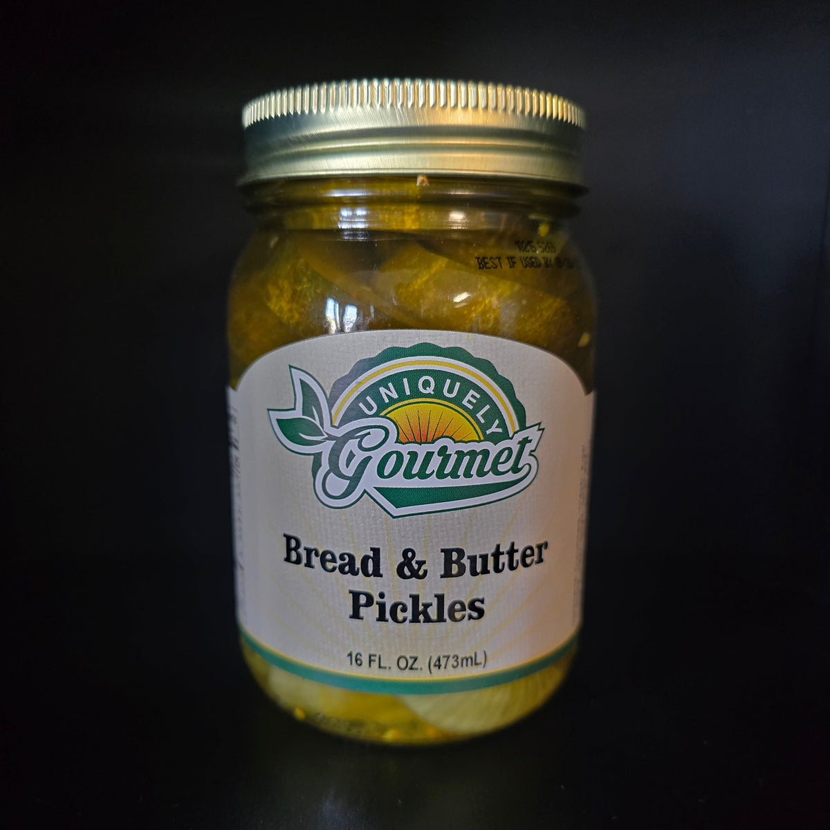 A glass jar of uniquely gourmet Bread & Butter Pickles with a white label and gold metal lid, set against a dark black background.