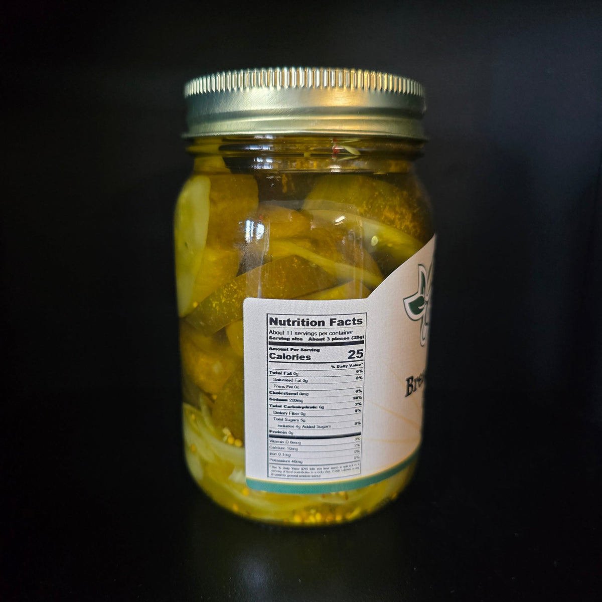 A glass mason jar filled with pickled yellow vegetables in oil, sealed with a silver metal lid and featuring a white label with nutrition facts, set against a dark black background.