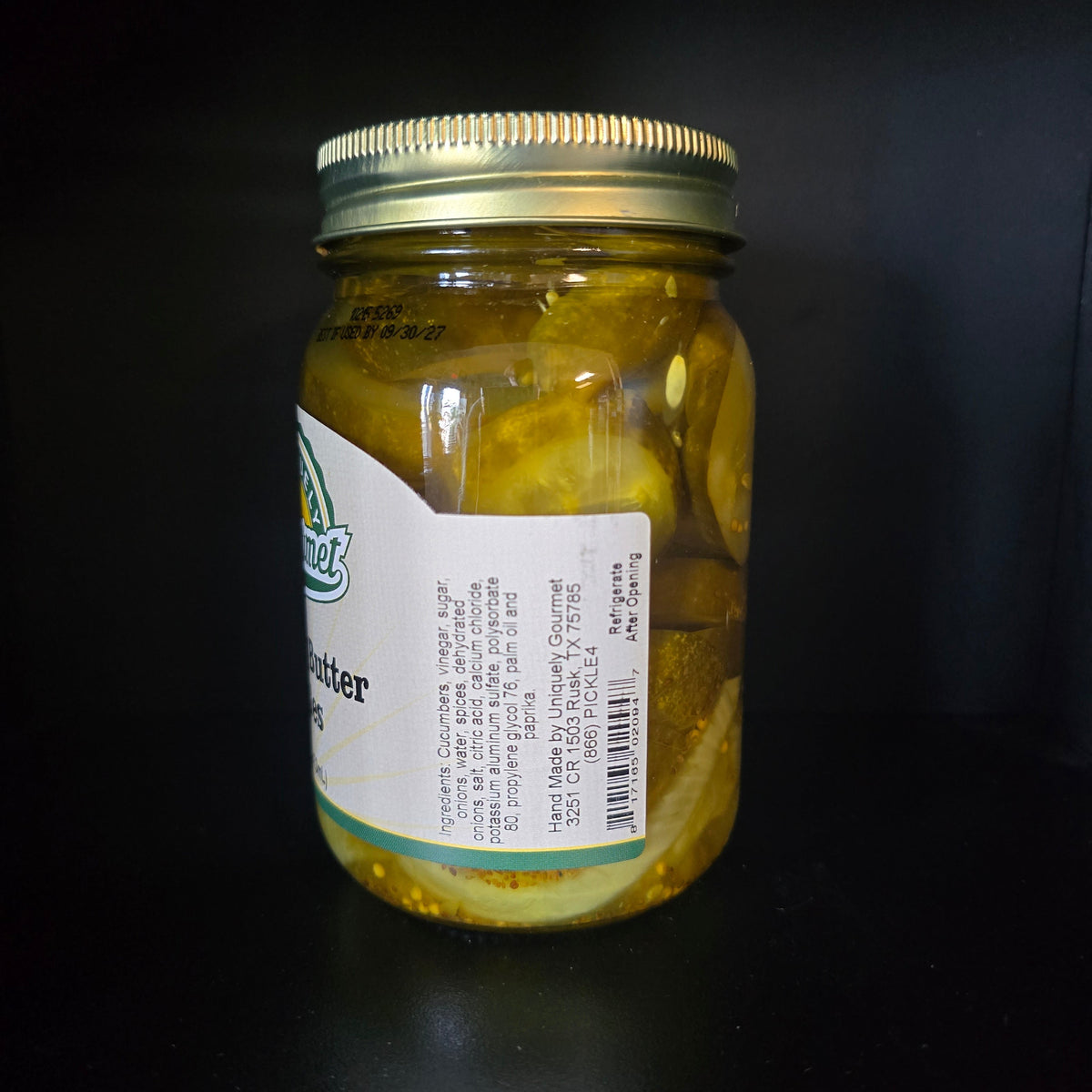 A glass jar of sliced pickles with a gold metal lid and a white label, photographed on a dark black background.