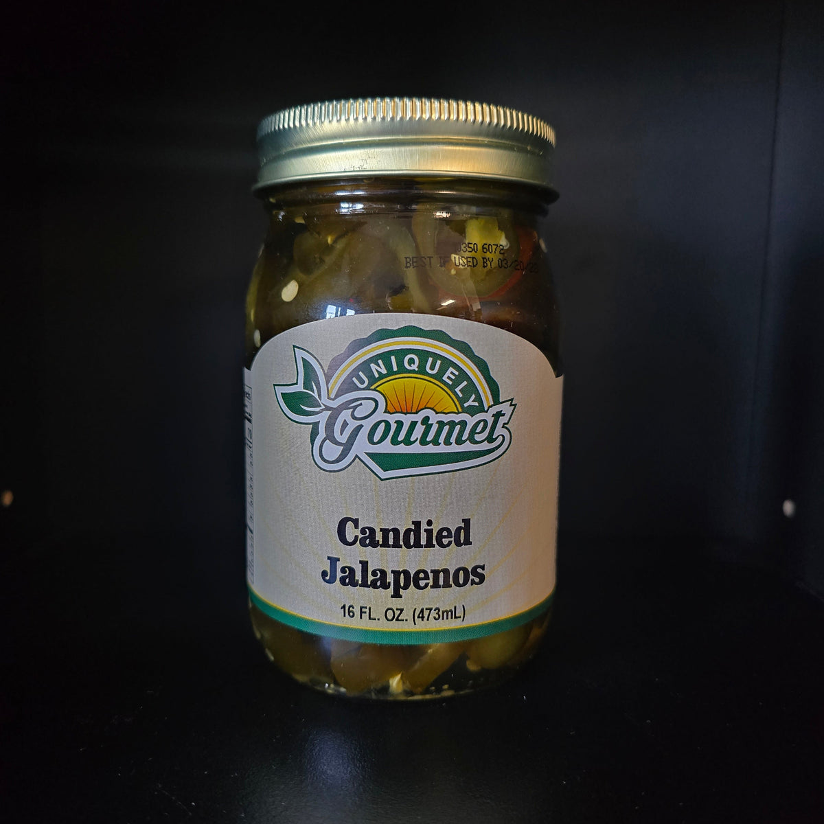 A glass jar of Unique Gourmet Candied Jalapeños with a white label and gold metal lid, set against a dark background.