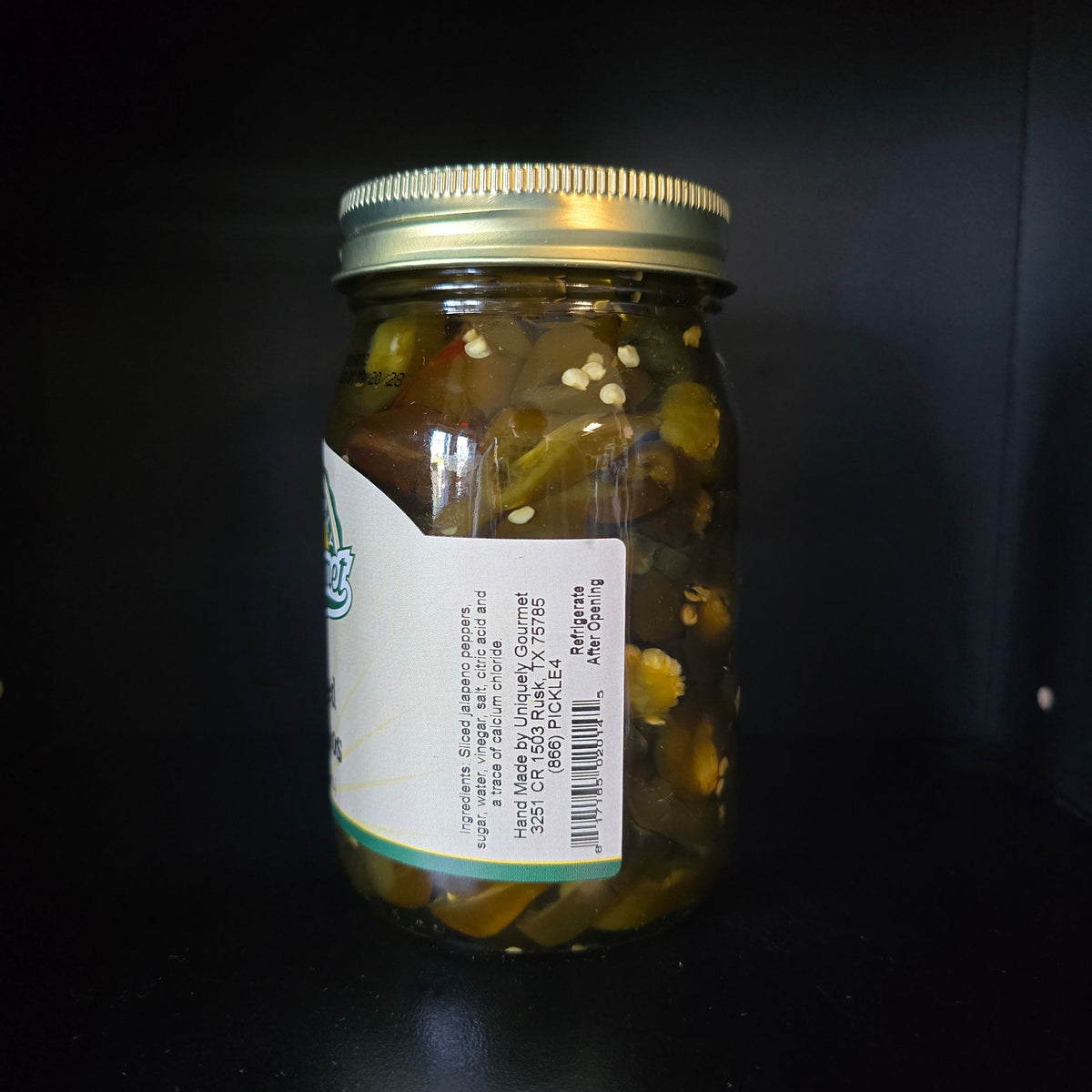 A glass jar of pickled vegetables with a white label and a gold metal lid, photographed against a dark black background.