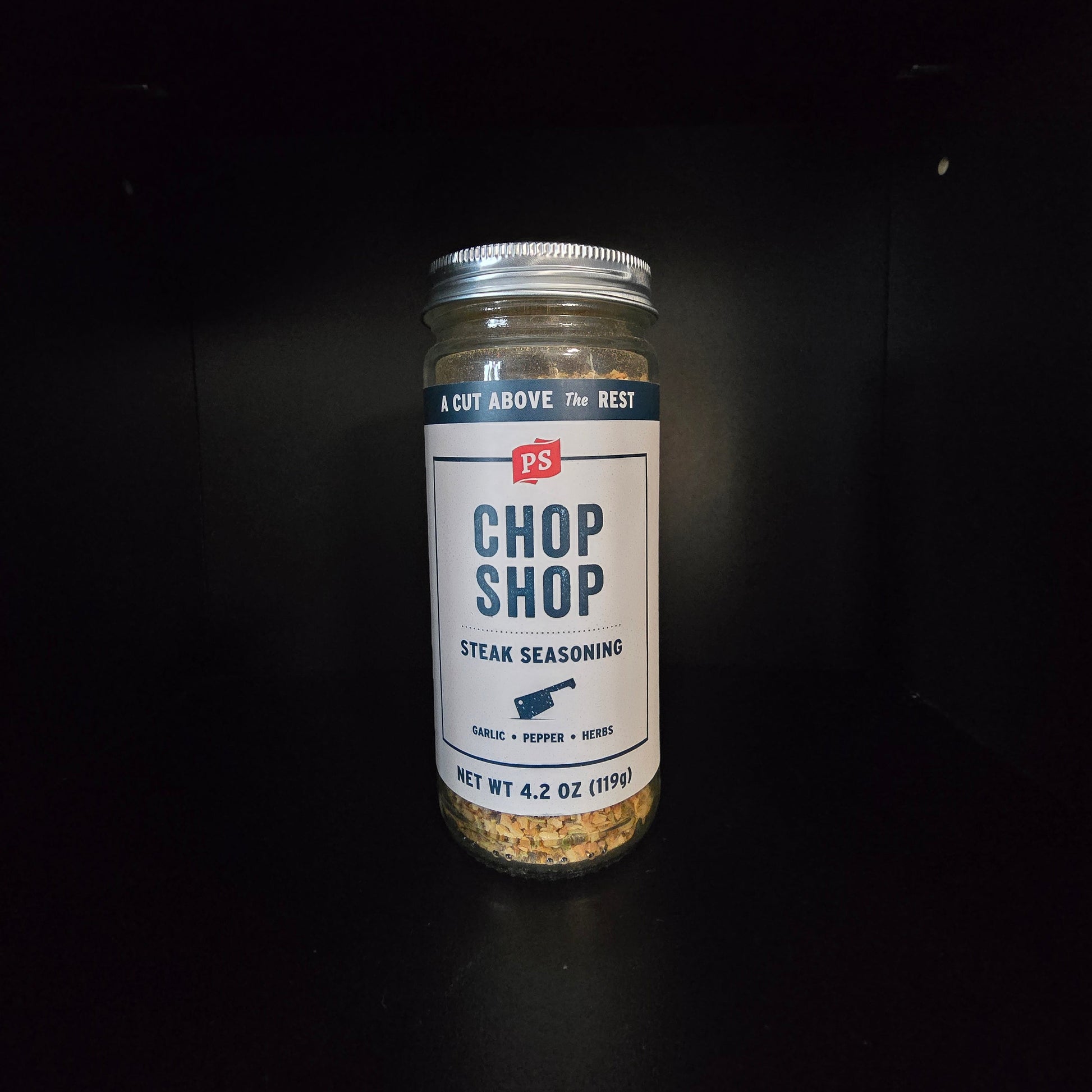 A glass jar of Chop Shop Steak Seasoning with a white label and silver lid, set against a dark black background.
