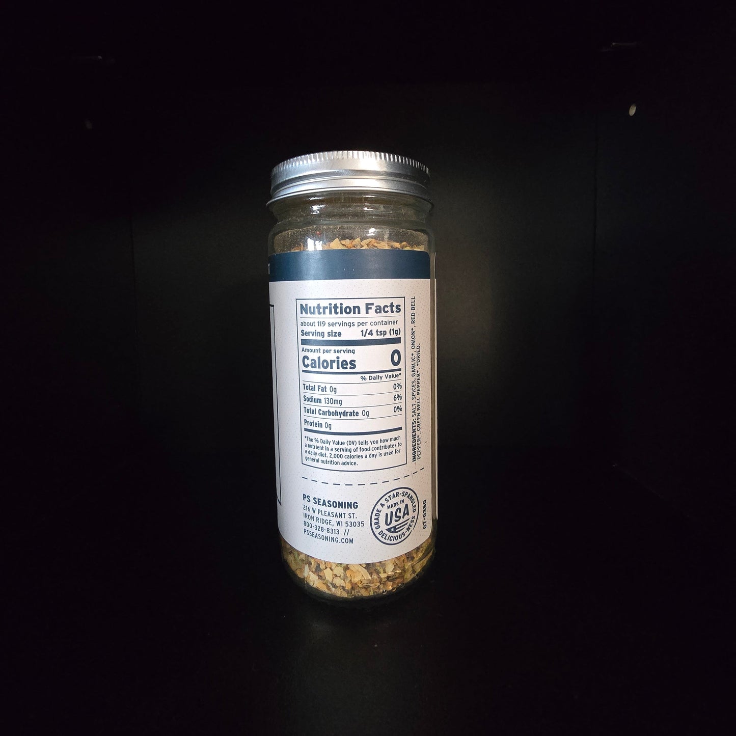 A clear glass jar filled with a beige and brown textured seasoning mix, featuring a white label with a blue stripe and a silver metal lid, set against a solid black background.