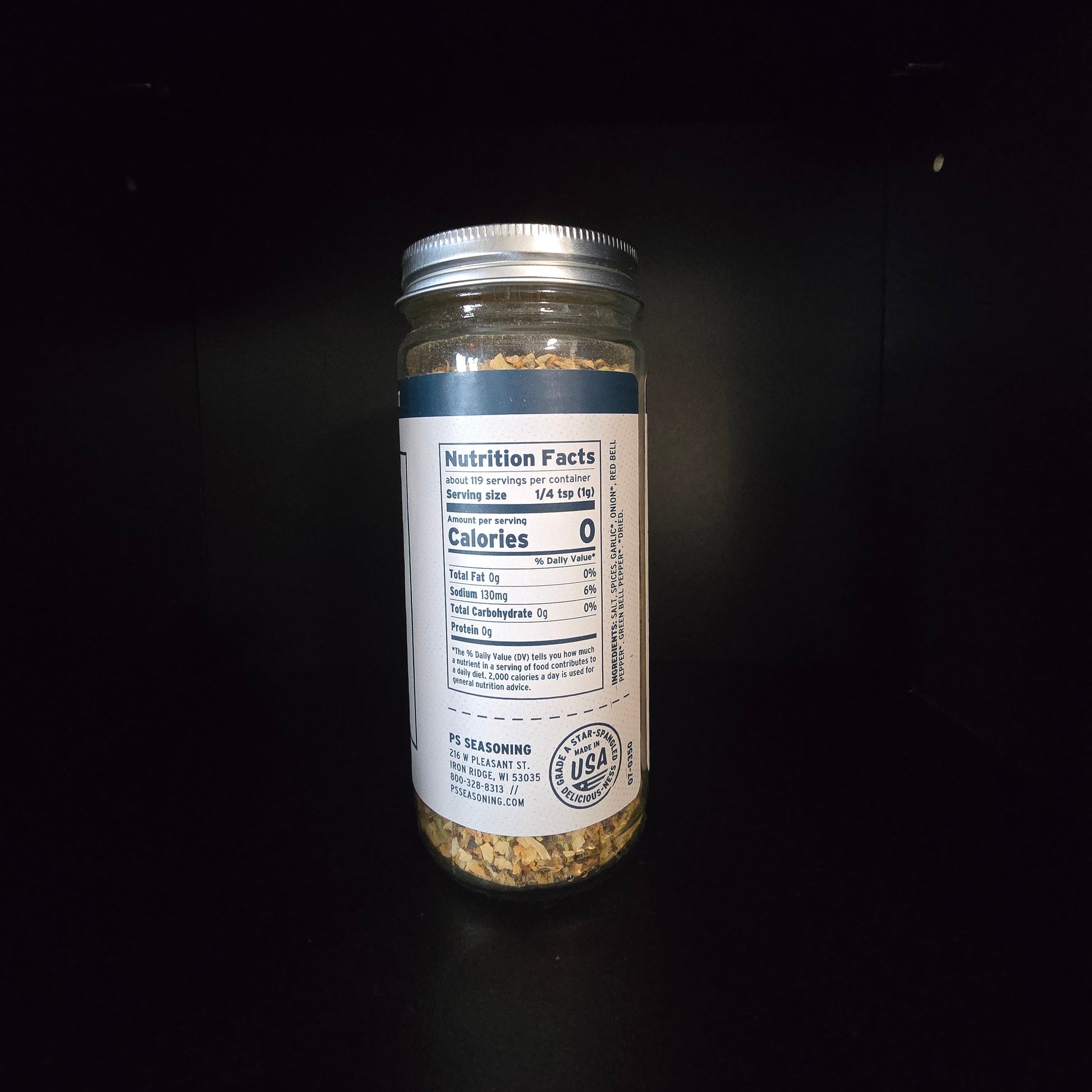 A clear glass jar filled with a beige and brown textured seasoning mix, featuring a white label with a blue stripe and a silver metal lid, set against a solid black background.