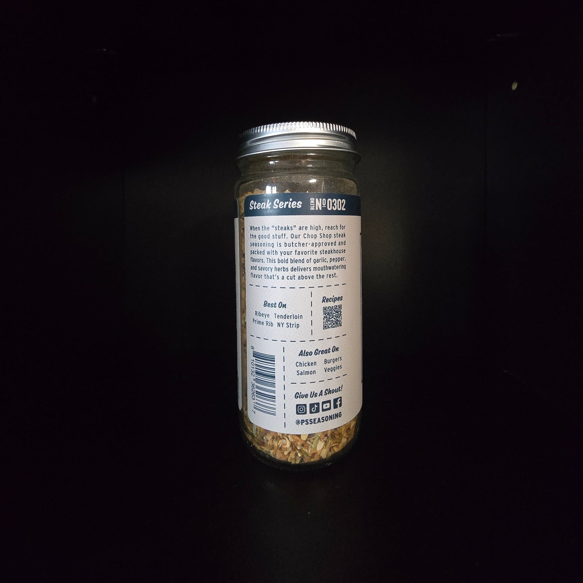 A glass jar of seasoning, showing the back label and silver lid against a dark black background.