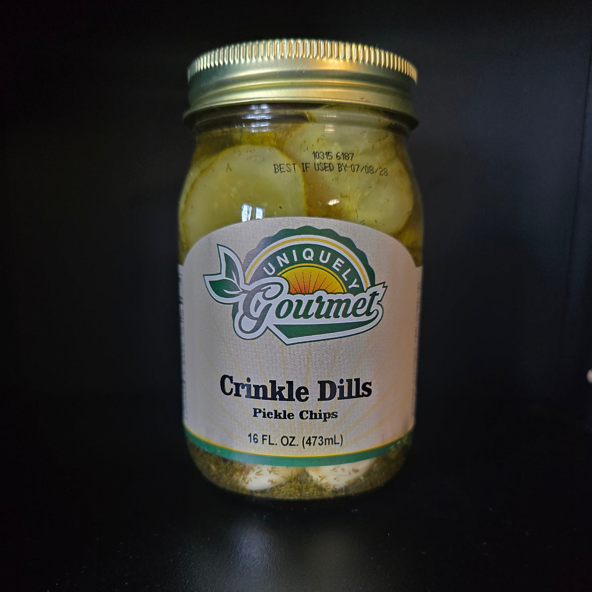 A glass jar of Uniquely Gourmet Crinkle Dills pickle chips with a gold metal lid and a white label, set against a dark black background.