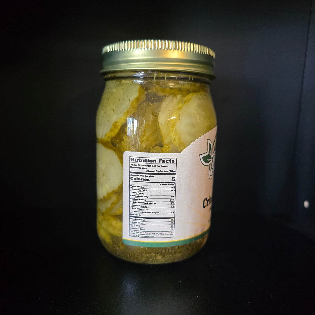 A glass jar filled with pickled or marinated vegetables, featuring a gold metal lid and a partially visible white label with green text, set against a dark black background.