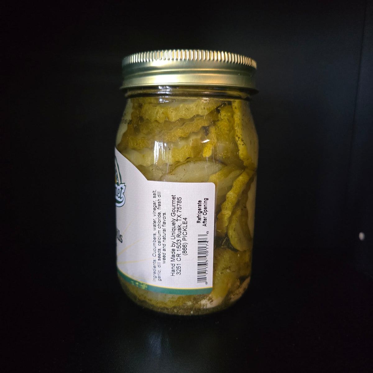 A glass jar of pickled cucumbers with a gold metal lid and a white label, set against a dark black background.