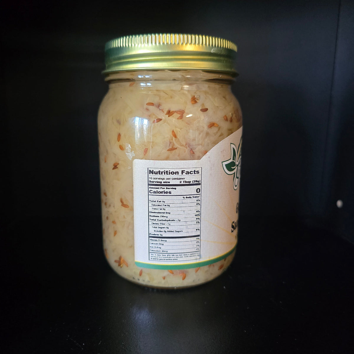 A glass jar of shredded pale beige food with visible orange flecks, sealed with a gold metal lid and a white label showing nutrition facts, photographed on a dark background.