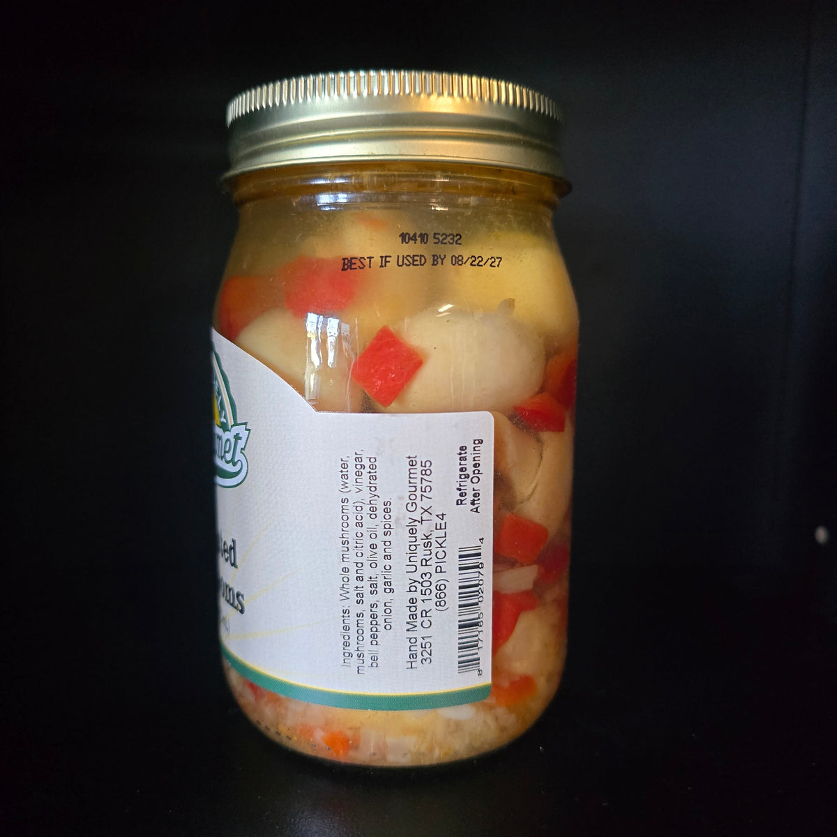 A glass jar of pickled onions with visible red and pale yellow vegetables in a clear liquid, sealed with a gold metal lid and featuring a white label, set against a dark background.