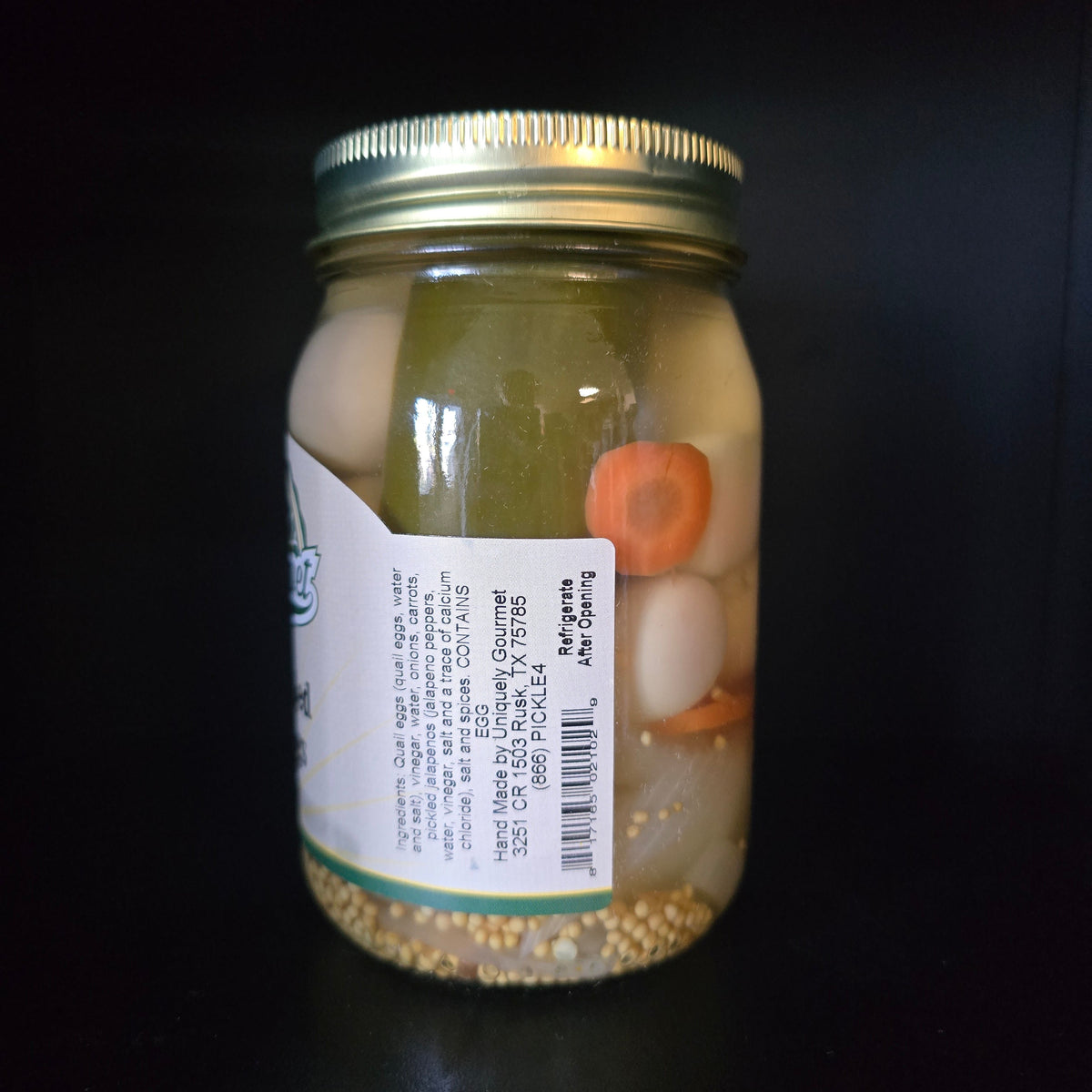 A glass mason jar filled with pickled onions and carrot slices, sealed with a gold metal lid and featuring a white product label, set against a solid black background.