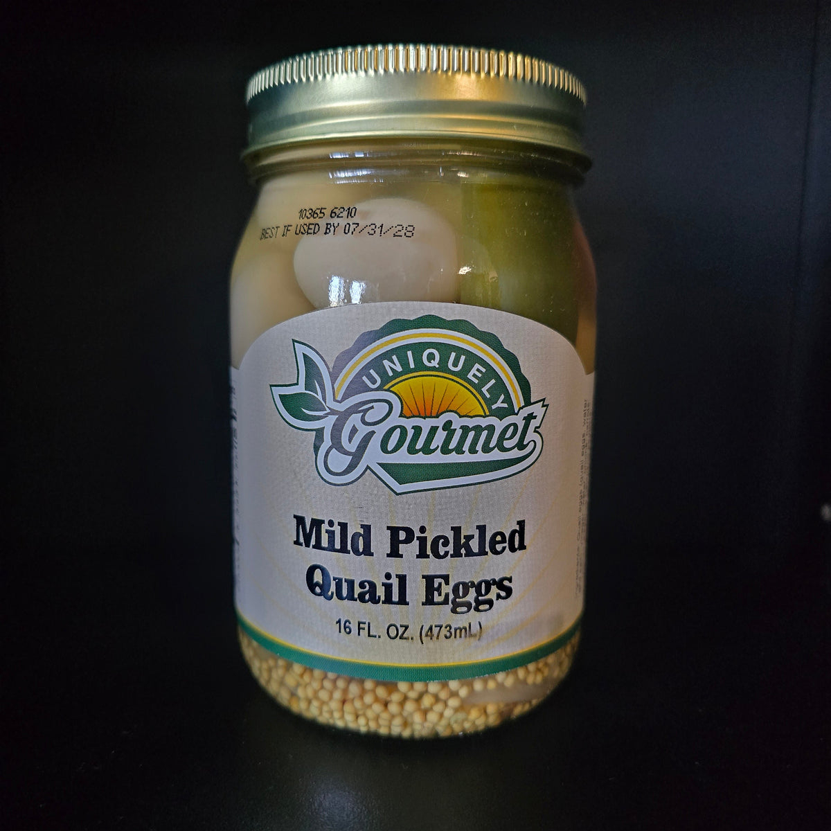 A jar of Mild Pickled Quail Eggs by Uniquely Gourmet, featuring a white label with green and gold accents and a gold metal lid, set against a dark background.