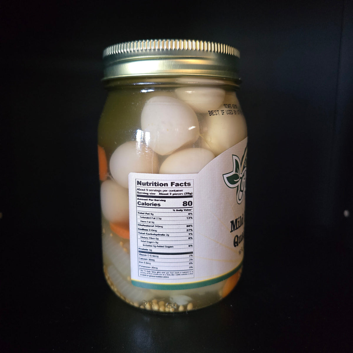 A glass jar filled with garlic onions in oil, sealed with a gold metal lid, and featuring a white label with nutrition facts, set against a dark background.