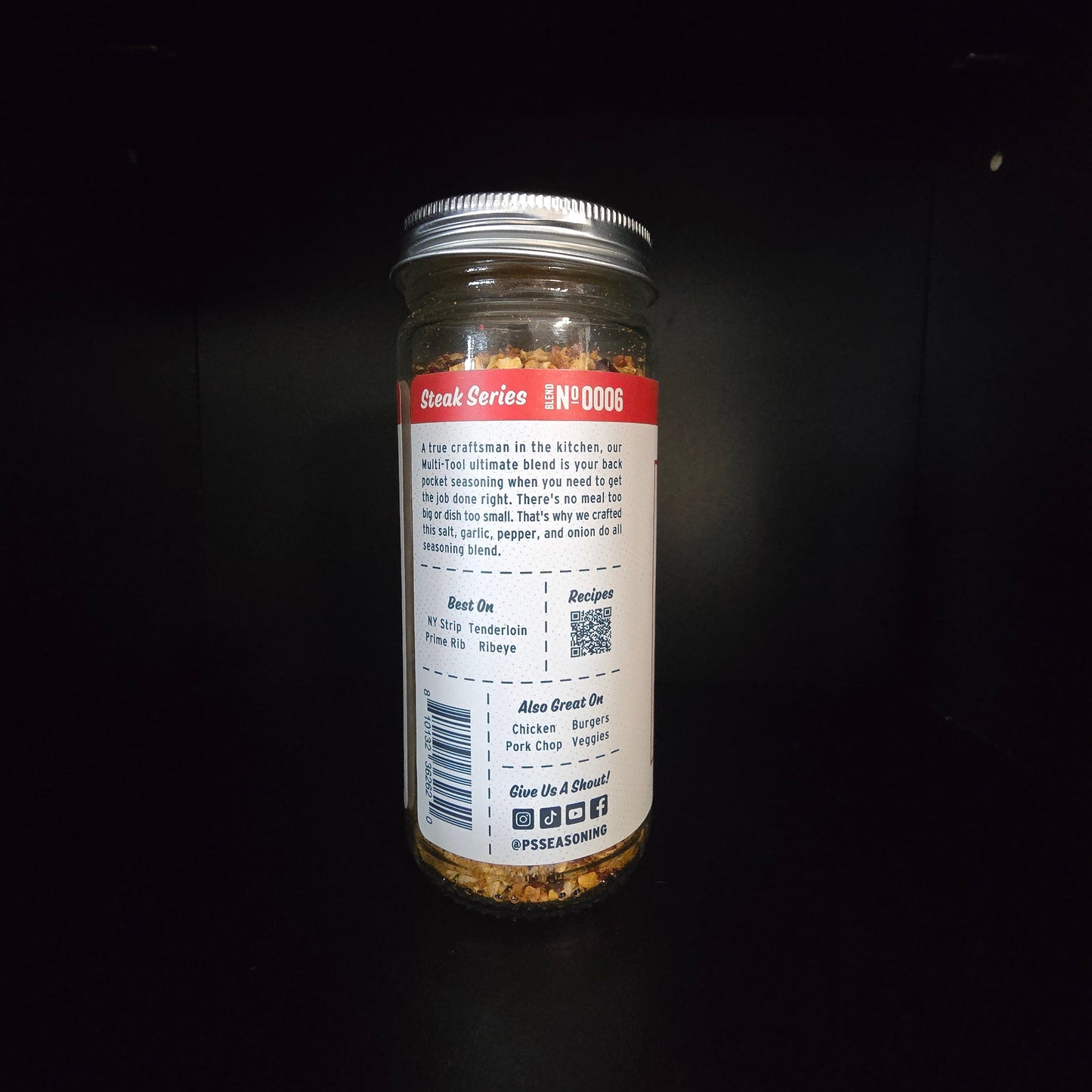 A glass spice jar with a silver metal lid, showing the back of the label with product details, set against a dark black background.