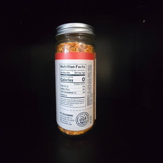 A clear glass jar filled with a reddish-brown seasoning blend, featuring a white label with a Nutrition Facts panel and a silver metal lid, set against a dark background.