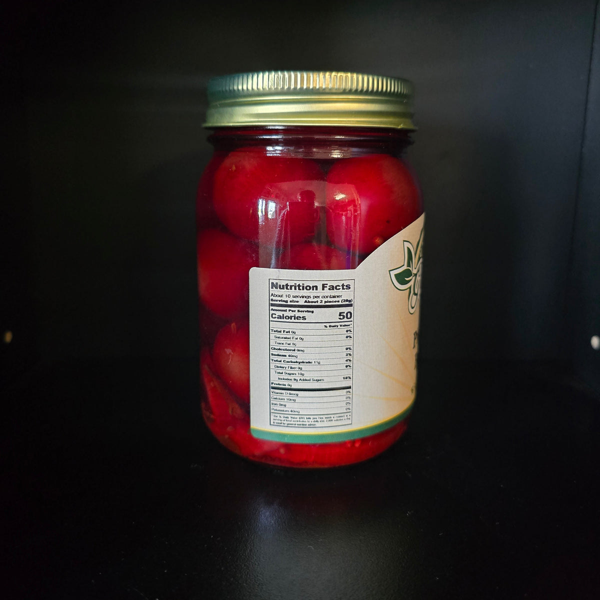 A glass jar filled with red pickled or preserved vegetables, sealed with a gold metal lid and featuring a partially visible cream label with green branding, set against a dark background.