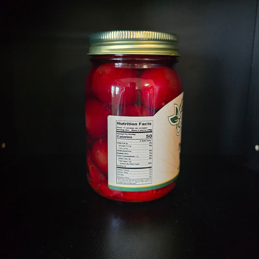 A glass jar filled with red pickled or preserved vegetables, sealed with a gold metal lid and featuring a partially visible cream label with green branding, set against a dark background.