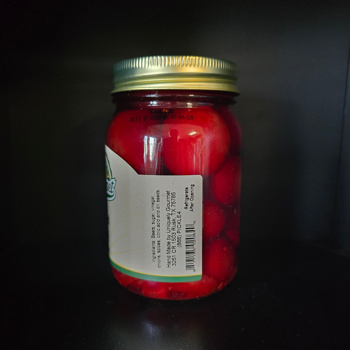 A glass jar filled with red fruit pieces, sealed with a gold metal lid and covered by a white label, set against a dark background.