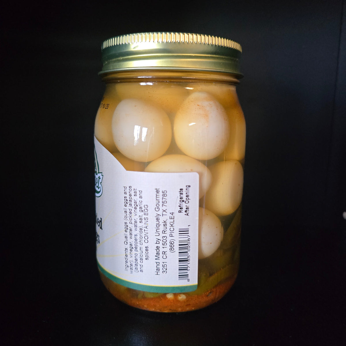 A glass jar filled with preserved eggs in oil, sealed with a gold metal lid and featuring a white label with text and a barcode, set against a solid black background.