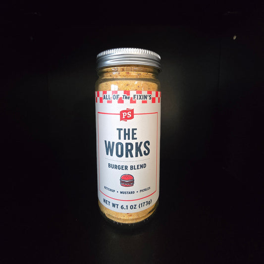 A glass jar of PS The Works Burger Blend seasoning mix on a solid black background. The jar has a white label with red and blue text, a silver metal lid, and the seasoning blend is visible through the clear glass.