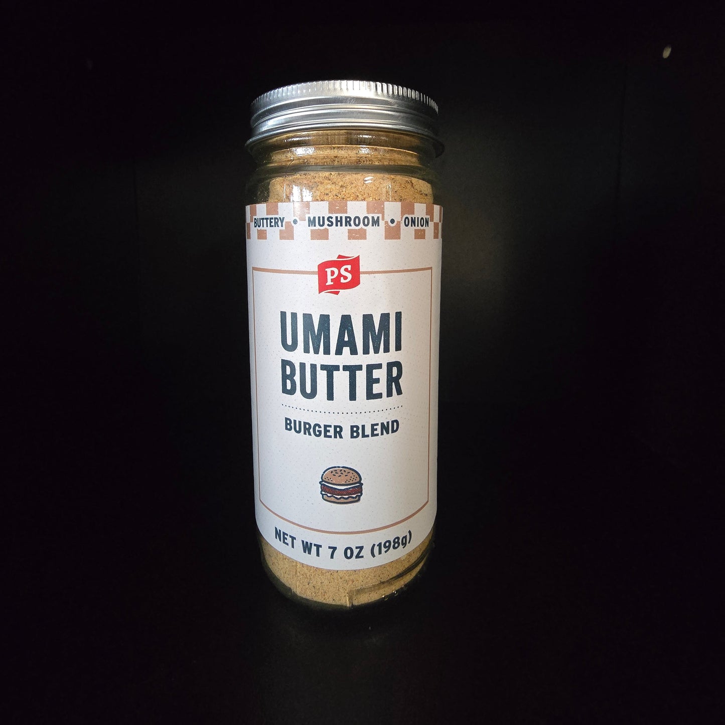 A glass jar of PS Umami Butter Burger Blend seasoning mix with a white label and silver lid, set against a solid black background.